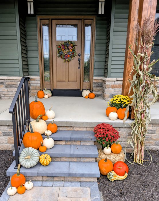 Fall pumpkins, mums and cornstalks arranged at a green porch and stone steps