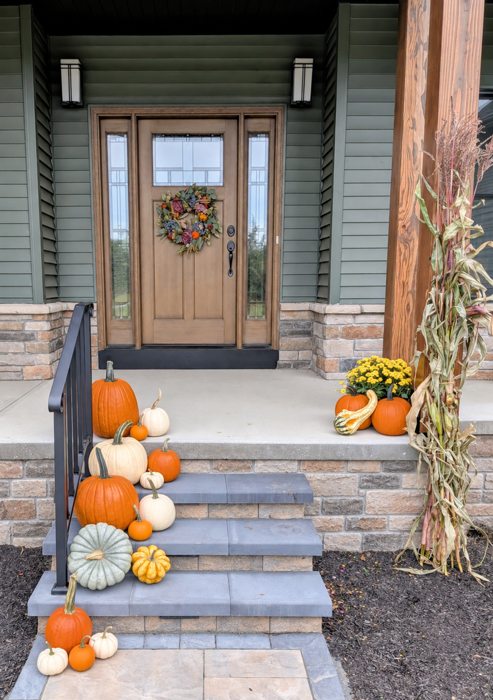 Fall pumpkins, mums and cornstalks arranged at a green porch and stone steps